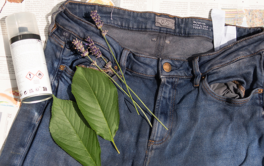 Eine alte Jeans wird mit einer Sprühdose besprüht. Auf der Hose liegen Blätter und Lavendel, darunter befindet sich eine Zeitung.